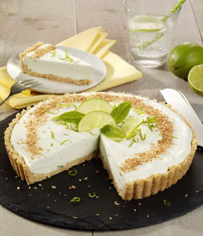 Limettentarte mit Limettenscheiben und Minzblättern, ein Stück auf einem Teller und Glas Wasser mit Limettenscheiben