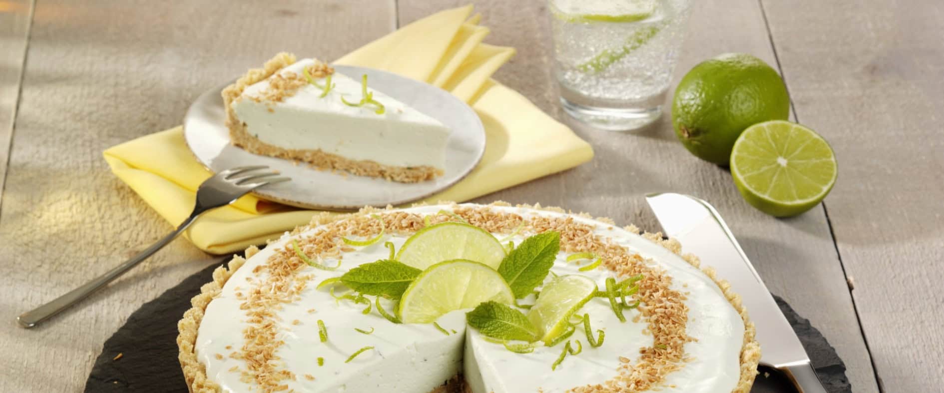 Limettentarte mit Limettenscheiben und Minzblättern, ein Stück auf einem Teller und Glas Wasser mit Limettenscheiben