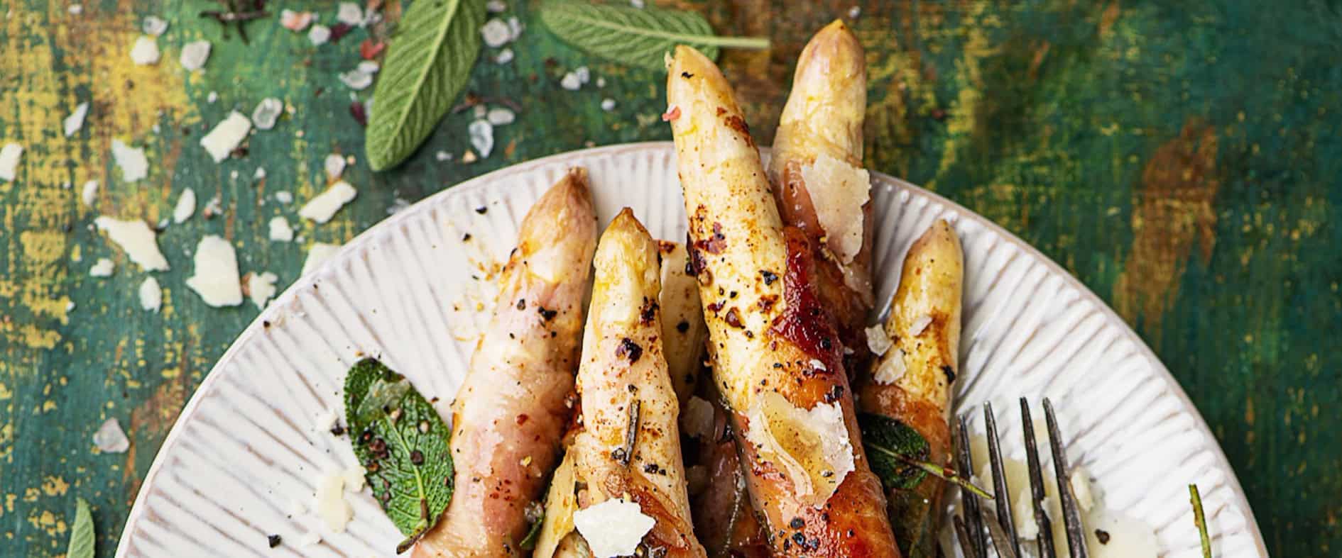 Saltimbocca vom weißen Spargel mit Prosciutto und Salbei auf einem Teller, garniert mit Parmesan und Salbeiblättern.