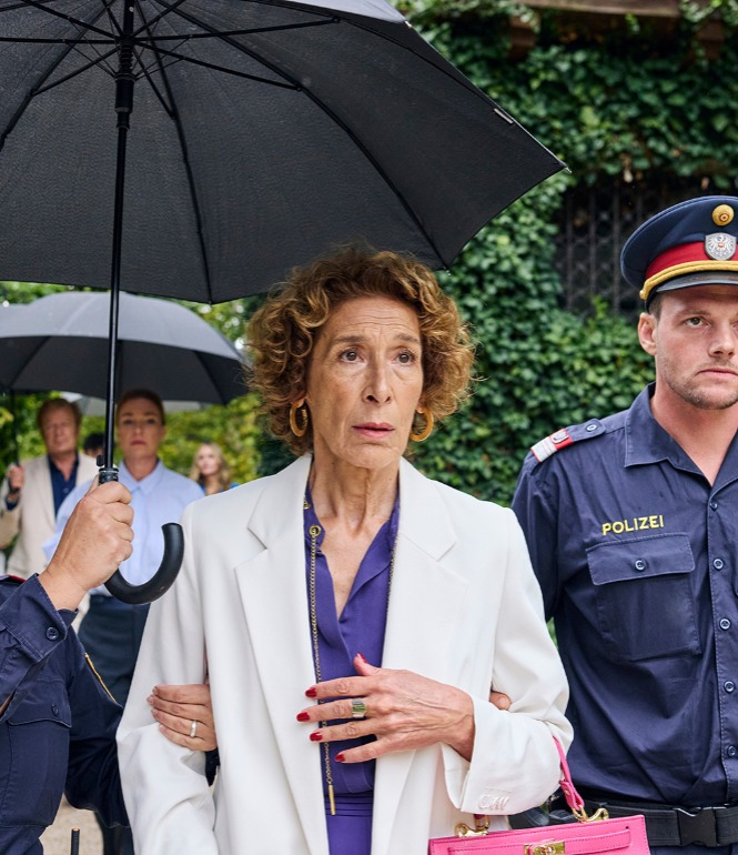 Gloria Almeda zwischen Polizisten, die linke Polizistin hält einen schwarzen Regenschirm über sie.