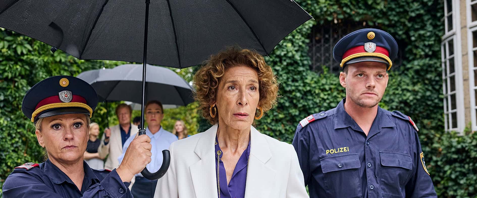 Gloria Almeda zwischen Polizisten, die linke Polizistin hält einen schwarzen Regenschirm über sie.