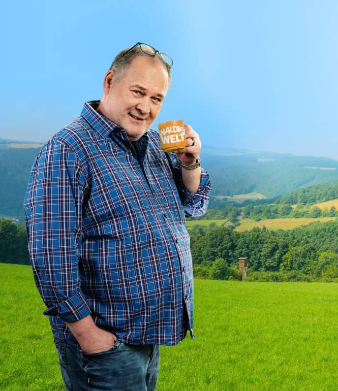 Walter Lehnertz hält eine Tasse mit der Aufschrift „Waldis Welt“ in einer grünen Landschaft