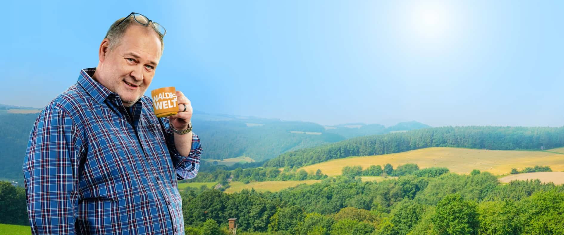 Walter Lehnertz hält eine Tasse mit der Aufschrift „Waldis Welt“ in einer grünen Landschaft