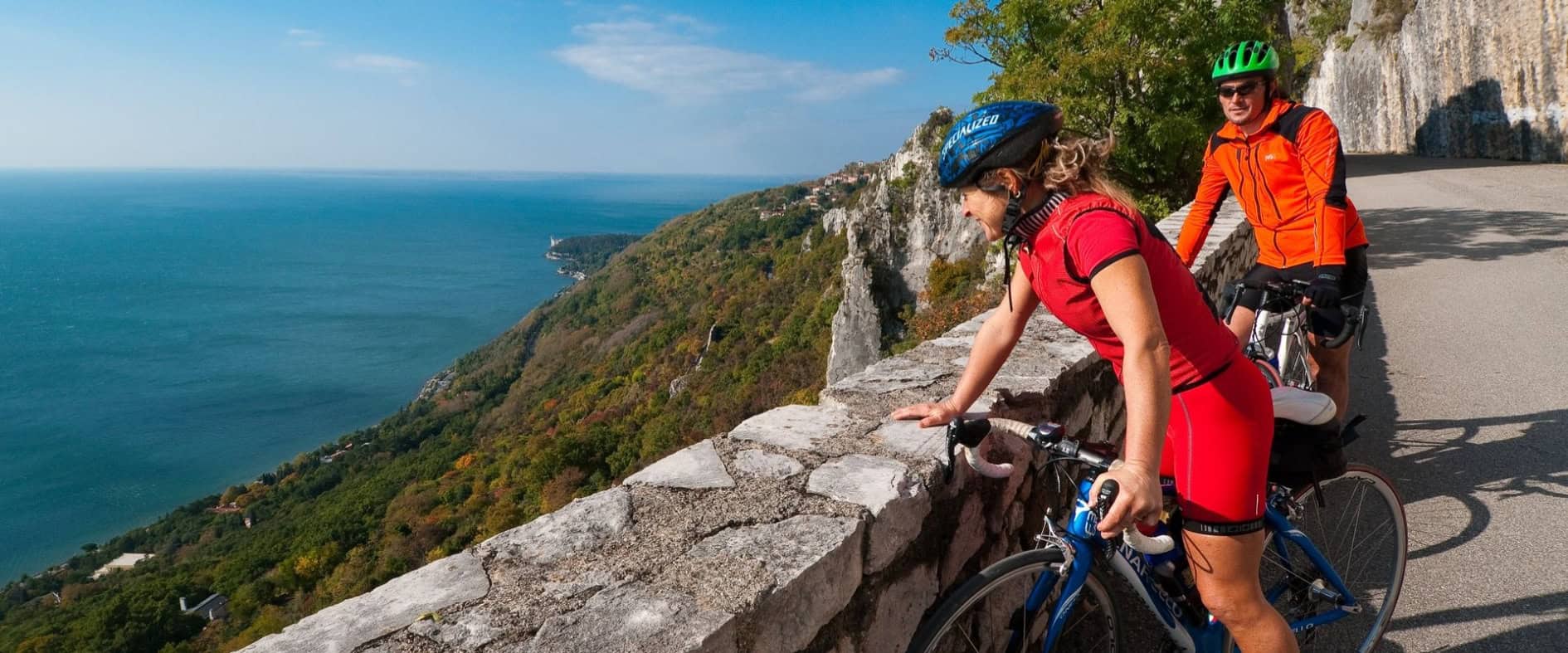 Zwei Personen mit Rennrädern machen eine Pause an einer steinernen Brüstung mit Blick auf das Meer und die Küstenlandschaft