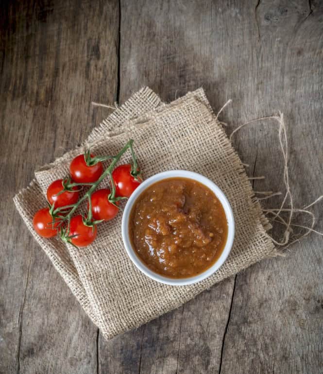 Schüssel mit fruchtig-würzigem Tomatenchutney, umgeben von frischen Kirschtomaten auf einem Jutesack und Holzuntergrund.