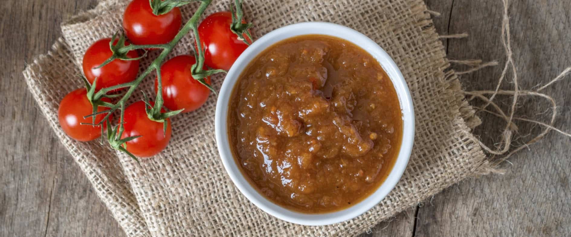 Schüssel mit fruchtig-würzigem Tomatenchutney, umgeben von frischen Kirschtomaten auf einem Jutesack und Holzuntergrund.