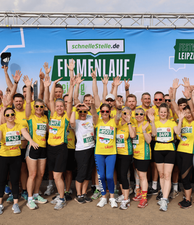 Große Gruppe von über 30 Teilnehmenden in gelben Laufshirts steht auf der Festwiese vor einer Eventwand mit „Firmenlauf Leipzig“-Schriftzug, viele haben die Arme gehoben und tragen Startnummern, im Hintergrund sind Sponsorentafeln und die Veranstaltungsarea zu sehen.