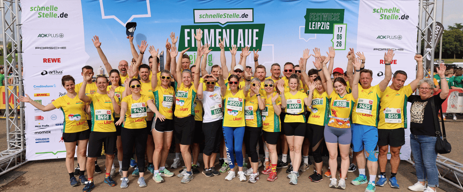 Große Gruppe von über 30 Teilnehmenden in gelben Laufshirts steht auf der Festwiese vor einer Eventwand mit „Firmenlauf Leipzig“-Schriftzug, viele haben die Arme gehoben und tragen Startnummern, im Hintergrund sind Sponsorentafeln und die Veranstaltungsarea zu sehen.