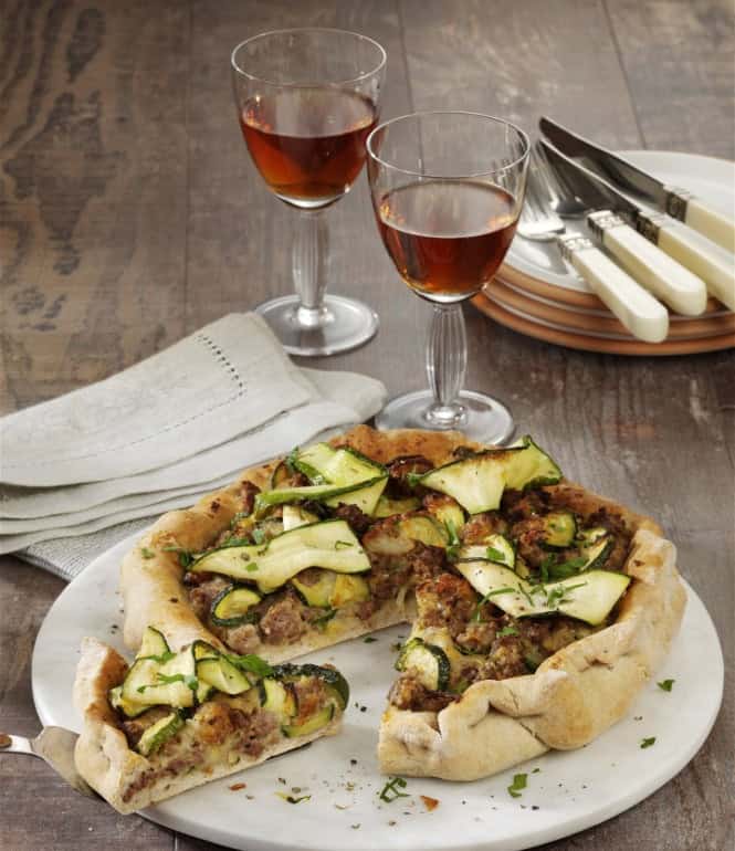 Hackfleisch-Zucchini-Quiche auf einem weißen Teller, serviert mit Rotwein und Besteck auf einem rustikalen Holztisch.