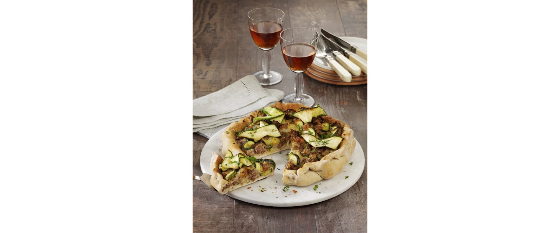Hackfleisch-Zucchini-Quiche auf einem weißen Teller, serviert mit Rotwein und Besteck auf einem rustikalen Holztisch.
