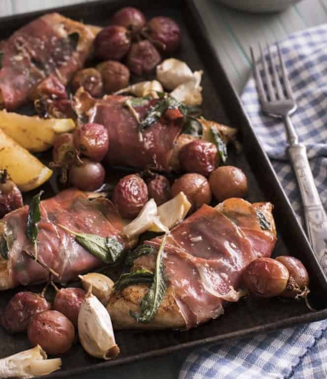 Hähnchen-Saltimbocca mit Parmaschinken, Salbei, gebratenen Trauben und Knoblauchzehen auf einem Backblech.