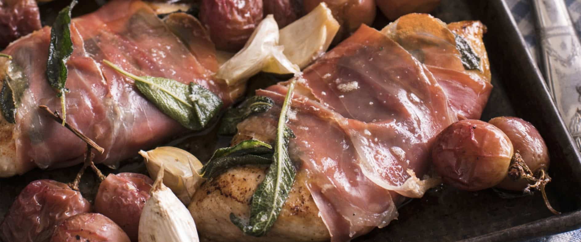 Hähnchen-Saltimbocca mit Parmaschinken, Salbei, gebratenen Trauben und Knoblauchzehen auf einem Backblech.