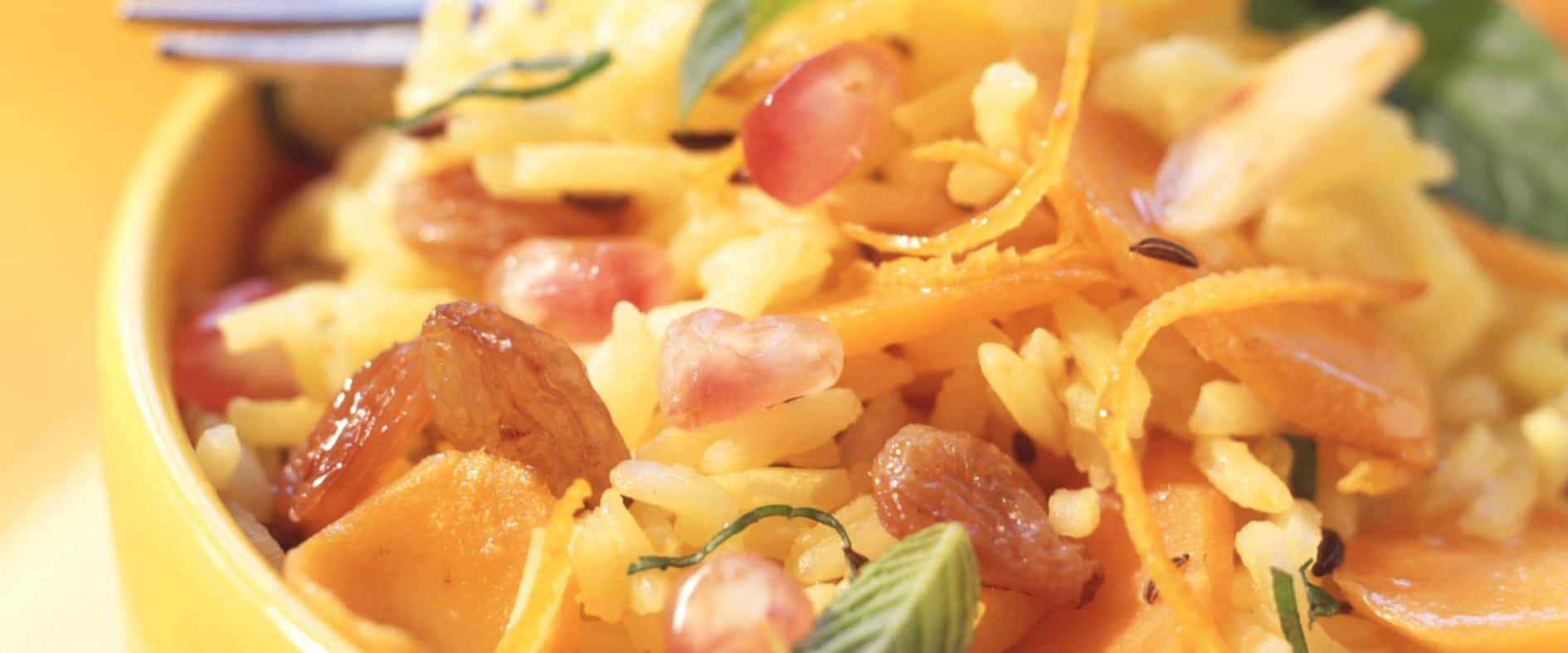 Indischer Reissalat mit Orangendressing, Granatapfelkernen, Rosinen, Möhren und Mandelstiften in einer gelben Schüssel, garniert mit Minze.