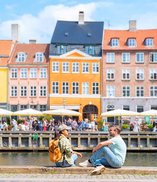 Zwei Personen genießen die entspannte Atmosphäre am Wasser in Kopenhagen mit Blick auf die bunten Häuser der Stadt und das lebhafte Treiben in den Cafés.