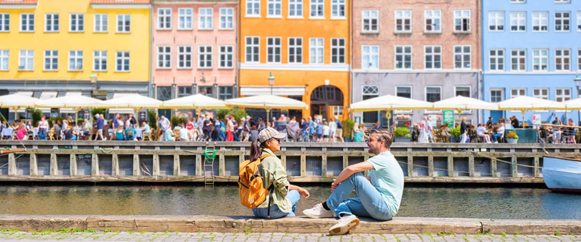 Zwei Personen genießen die entspannte Atmosphäre am Wasser in Kopenhagen mit Blick auf die bunten Häuser der Stadt und das lebhafte Treiben in den Cafés.