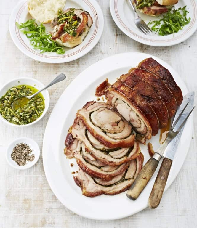 Knusprige Porchetta mit grüner Salsa in Scheiben geschnitten auf einem weißen Teller, serviert mit Brötchen, Rucola und einer Schüssel grüner Salsa.