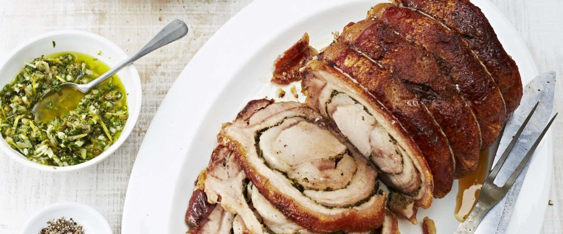 Knusprige Porchetta mit grüner Salsa in Scheiben geschnitten auf einem weißen Teller, serviert mit Brötchen, Rucola und einer Schüssel grüner Salsa.