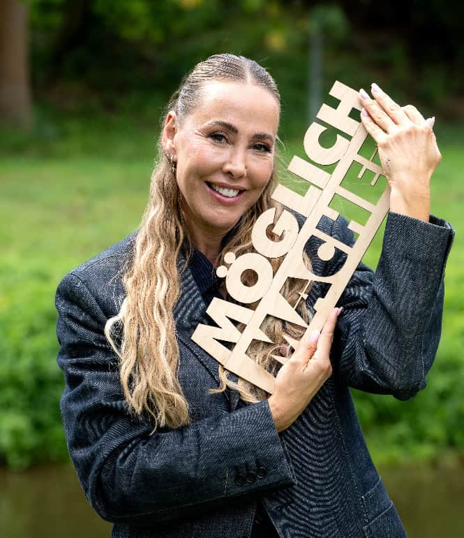 Sylvia Walker präsentiert den Sachsenlotto-Möglichmacher mit Holzschild als Symbol für Engagement, Unterstützung und die vielfältige Förderkampagne in Sachsen