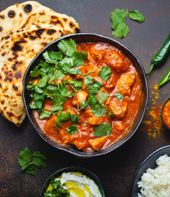 Traditionelles indisches Curry mit frischem Koriander, serviert in einer Schüssel, umgeben von Naan-Brot, Basmatireis, Gewürzen und frischen Kräutern.