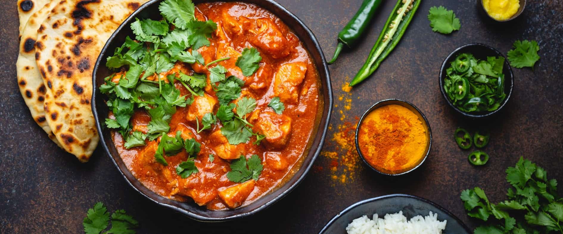 Traditionelles indisches Curry mit frischem Koriander, serviert in einer Schüssel, umgeben von Naan-Brot, Basmatireis, Gewürzen und frischen Kräutern.