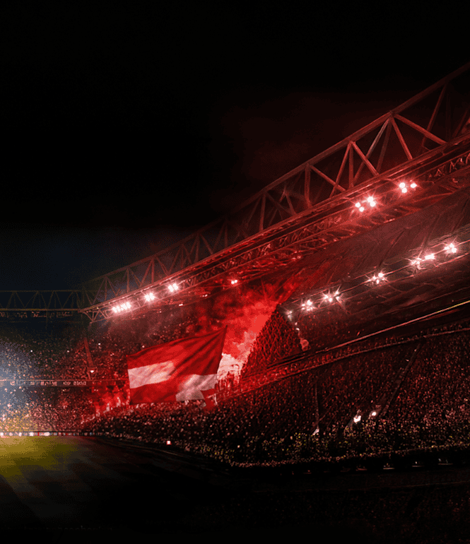 Fußball-Fans mit gelben und roten Fahnen, Rauch und Flutlicht sorgen im Stadion für eine elektrisierende Atmosphäre.