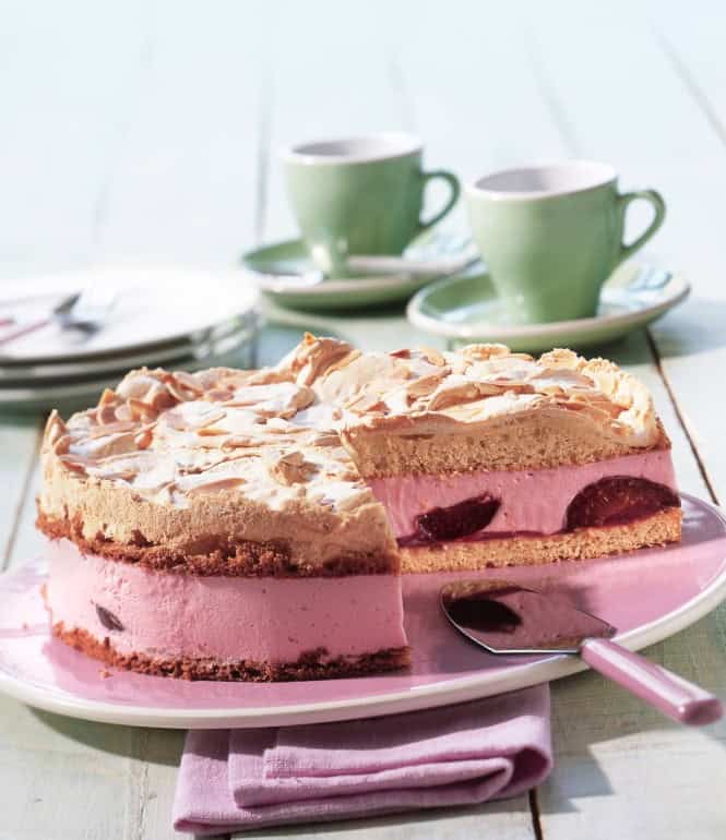 Pflaumen-Baisertorte mit knusprigem Baiserboden und Mandelblättchen, auf einem Tortenteller mit einer aufgeschnittenen Scheibe, serviert mit frischen Pflaumen.