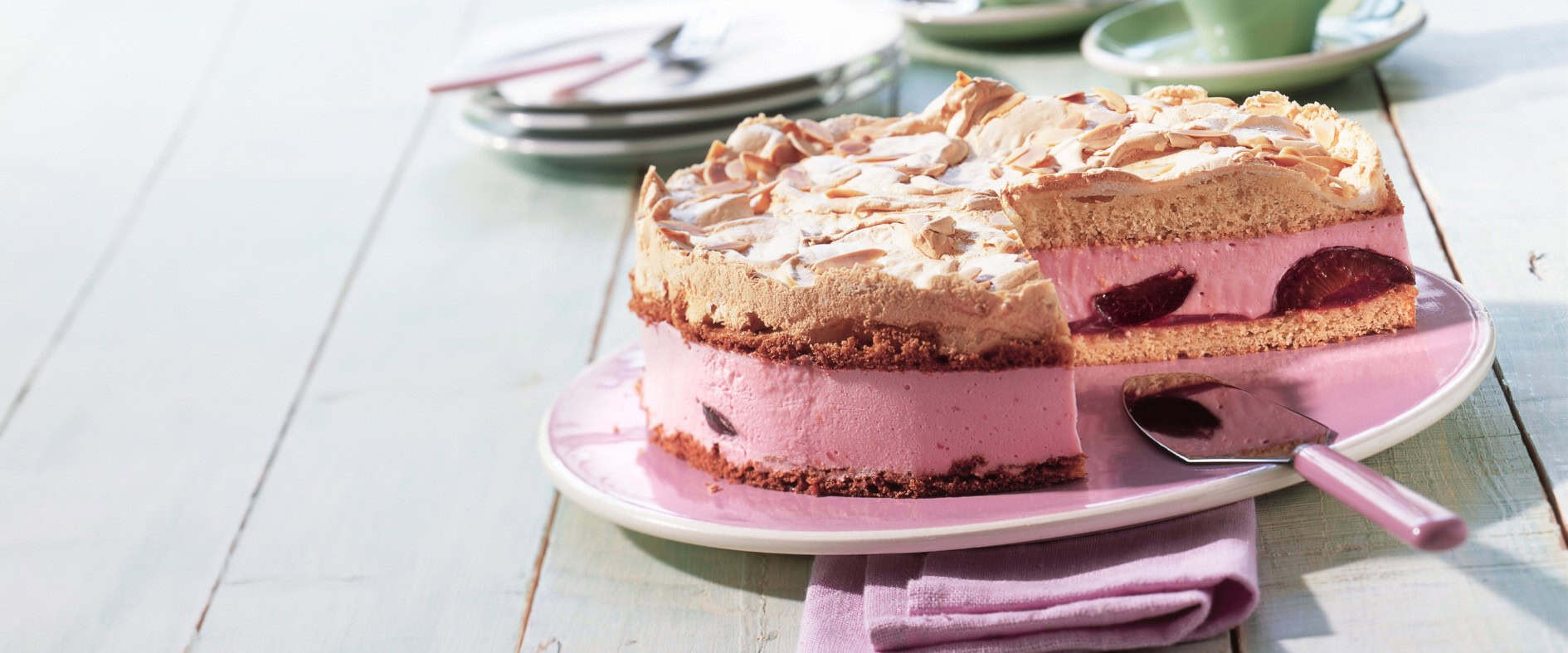 Pflaumen-Baisertorte mit knusprigem Baiserboden und Mandelblättchen, auf einem Tortenteller mit einer aufgeschnittenen Scheibe, serviert mit frischen Pflaumen.