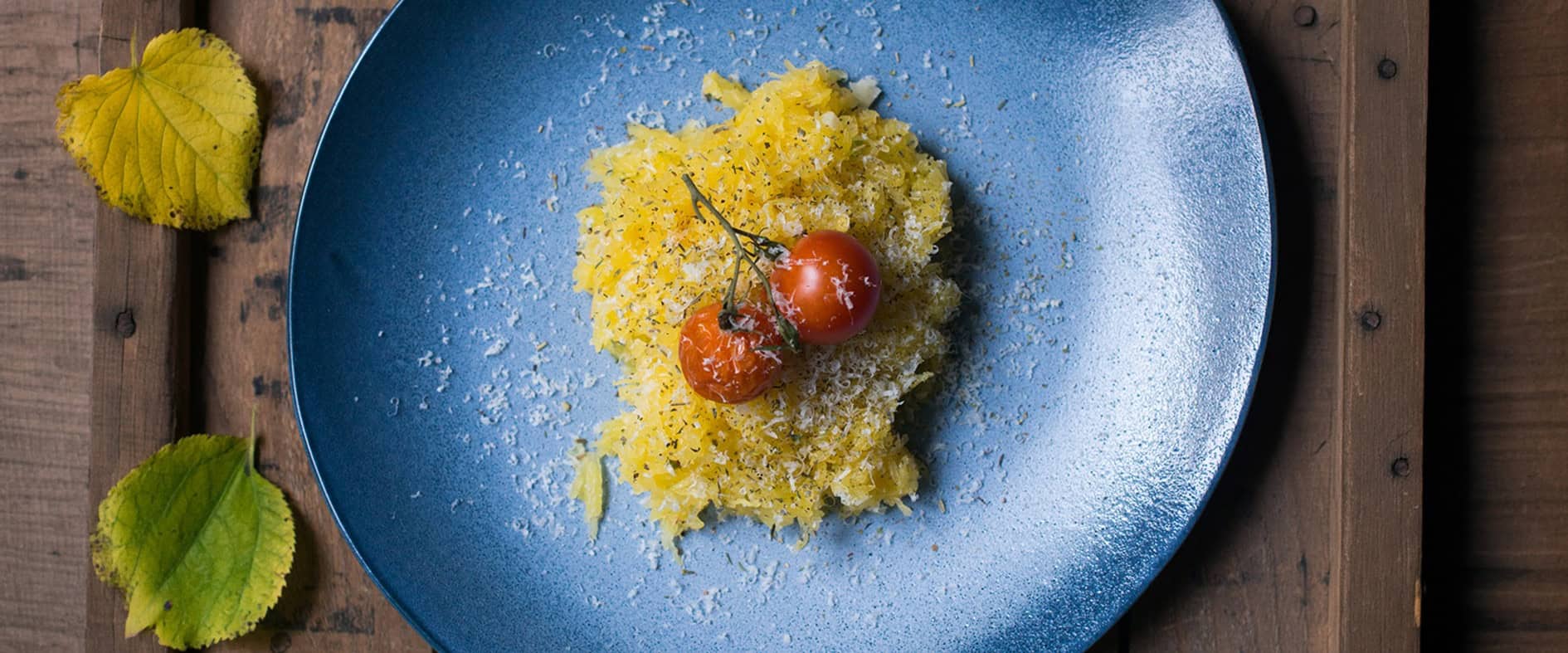 Teller mit Spaghettikürbis als Pasta-Alternative, garniert mit Käse und Tomate, serviert auf Holztisch mit Herbstblättern