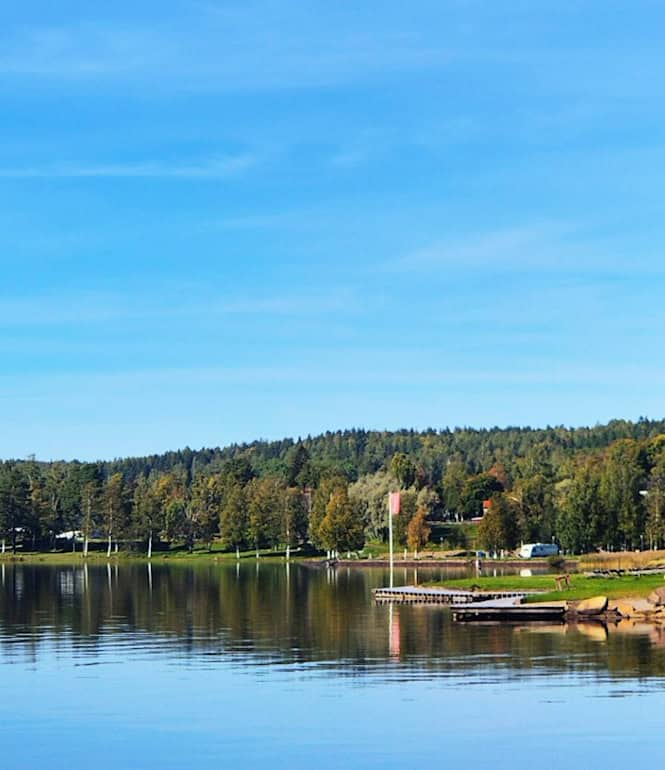 Idyllische Landschaft am Siljansee in Dalarna, Schweden: Eine weiße Kirche mit Turm am Ufer des ruhigen Sees, umgeben von grünen Bäumen und klarem Himmel.