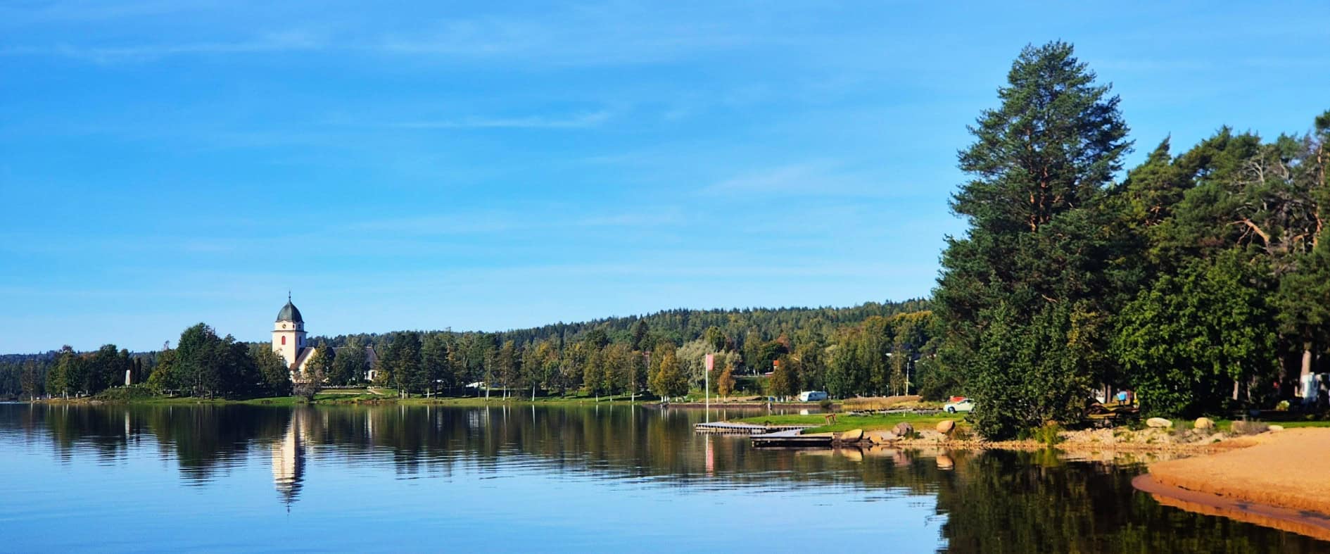 Idyllische Landschaft am Siljansee in Dalarna, Schweden: Eine weiße Kirche mit Turm am Ufer des ruhigen Sees, umgeben von grünen Bäumen und klarem Himmel.