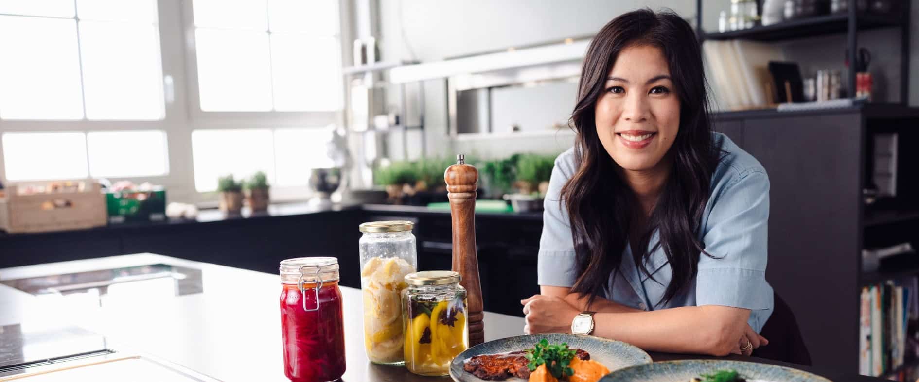 Mai Thi Nguyen-Kim lächelt in einer Küche und stützt sich auf eine Arbeitsplatte, vor ihr mehrere Teller mit Speisen und Gläser mit eingelegten Lebensmitteln
