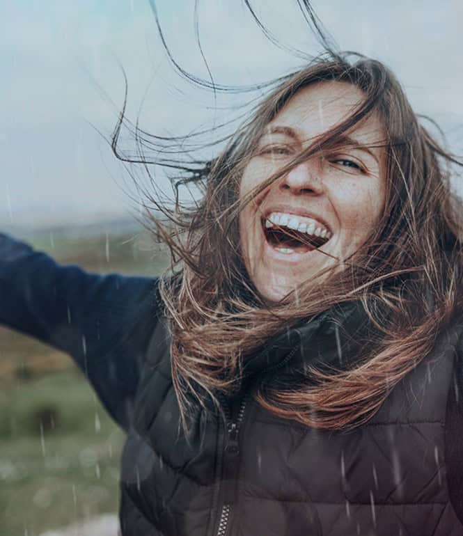 Lachende Frau im Freien mit ausgebreiteten Armen und im Wind wehendem Haar vor hügeliger, regnerischer Landschaft