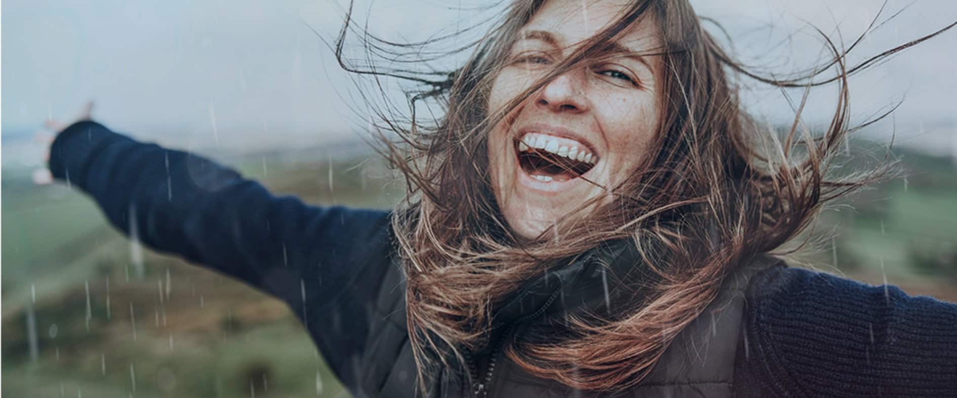 Lachende Frau im Freien mit ausgebreiteten Armen und im Wind wehendem Haar vor hügeliger, regnerischer Landschaft